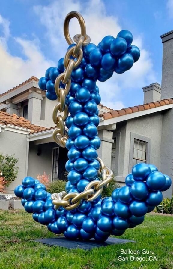 Anchor shaped sculpture made of blue balloons with a gold colored chain, placed in the front yard of a big house.