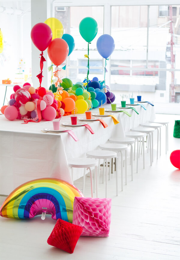 Rainbow colored balloon tablescape centerpiece with some single balloons floating above, set on a large white table with color matching cups.