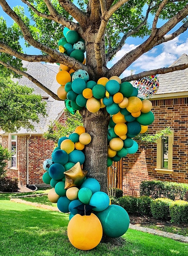 Balloon garland in teal, yellow and gold colors wrapped around a tree.